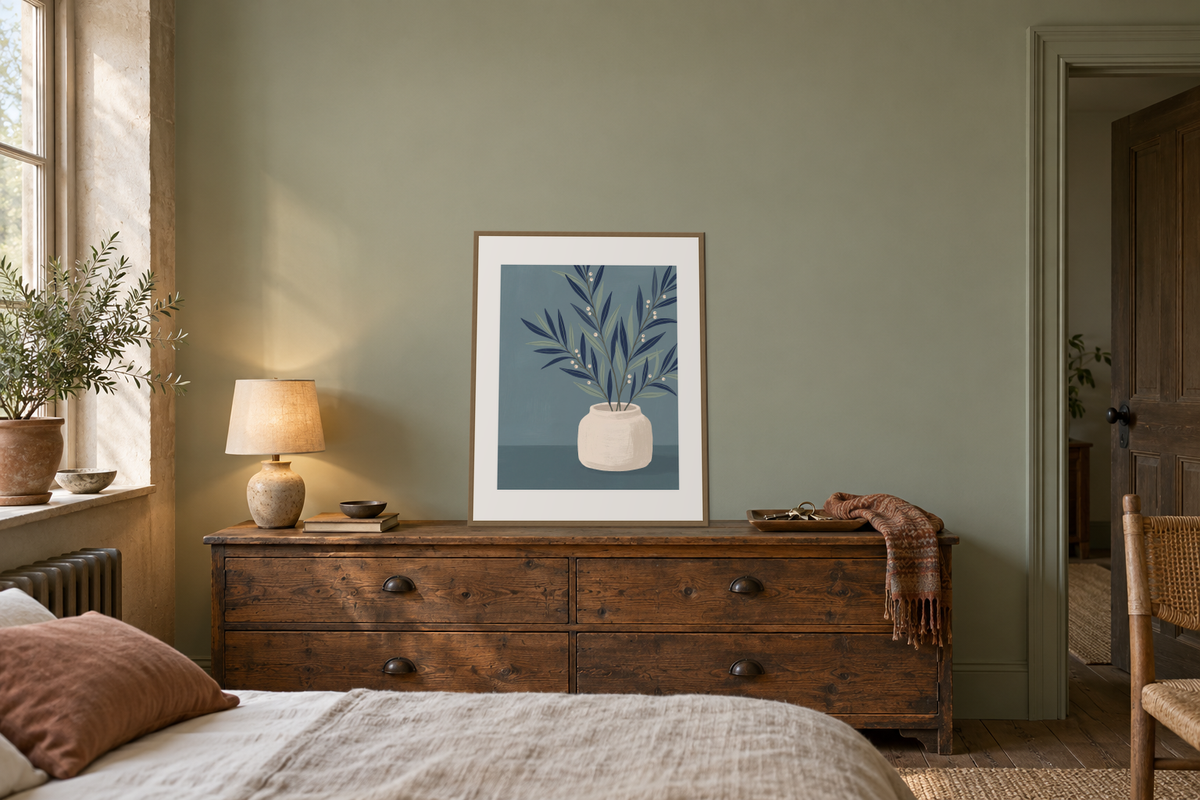 A framed botanical print leaning on a wooden dresser, with its full artwork visible between a lamp and a tray.